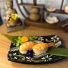 Two Nigiri Shrimp pieces are elegantly arranged on a decorative black plate adorned with green leaves and a small flower garnish, all set atop a wooden table. In the serene background, a Buddha statue and shelf evoke the ambiance of a sushi bistro in Edinburgh.