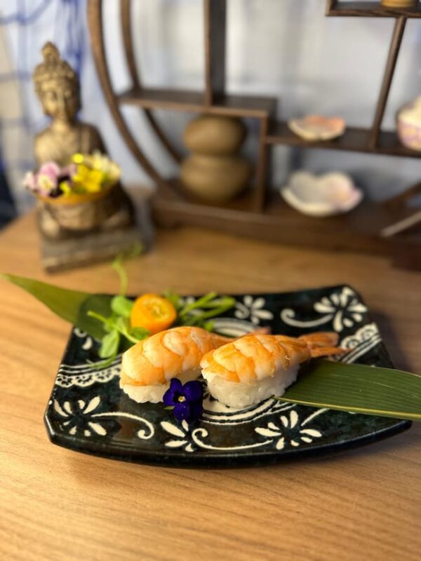 Two Nigiri Shrimp pieces are elegantly arranged on a decorative black plate adorned with green leaves and a small flower garnish, all set atop a wooden table. In the serene background, a Buddha statue and shelf evoke the ambiance of a sushi bistro in Edinburgh.