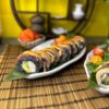 A delightful serving of "Phila with Black Tuna" sushi rolls is garnished with sauce and beautifully arranged on a bamboo mat against a yellow background, adorned with decorative items reminiscent of an Edinburgh sushi eatery.