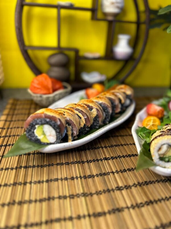 A delightful serving of "Phila with Black Tuna" sushi rolls is garnished with sauce and beautifully arranged on a bamboo mat against a yellow background, adorned with decorative items reminiscent of an Edinburgh sushi eatery.