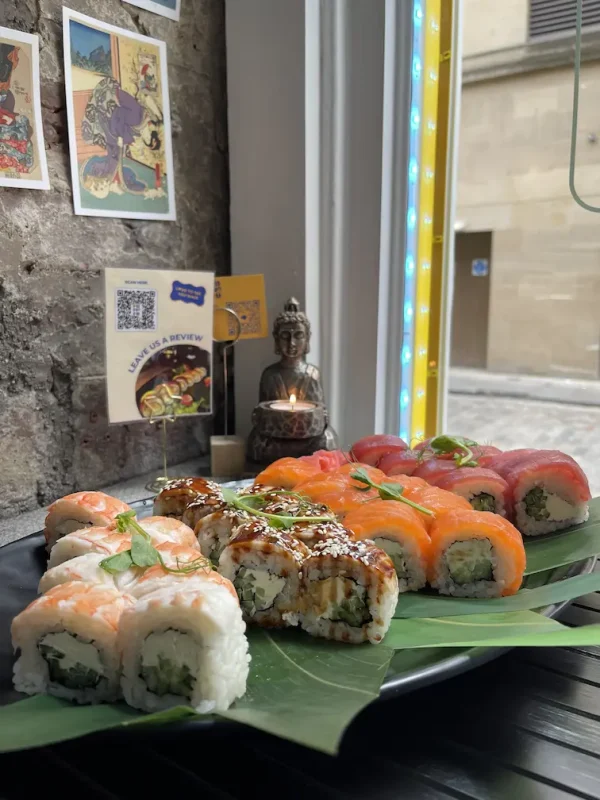 A platter from the Sushi Set 2 rests on a banana leaf, evoking the charm of an Edinburgh street market. In the background, a candle and Buddha statue accompany postcards on a brick wall, enhancing the serene ambiance.