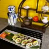 A dragon roll garnished with sesame seeds and wasabi on a black tray, next to decorative figurines and vases on a shelved display with a yellow background.