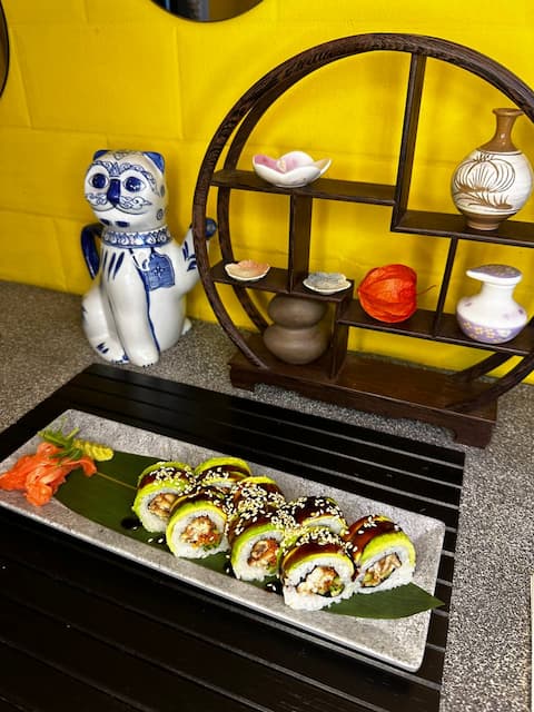 A dragon roll garnished with sesame seeds and wasabi on a black tray, next to decorative figurines and vases on a shelved display with a yellow background.