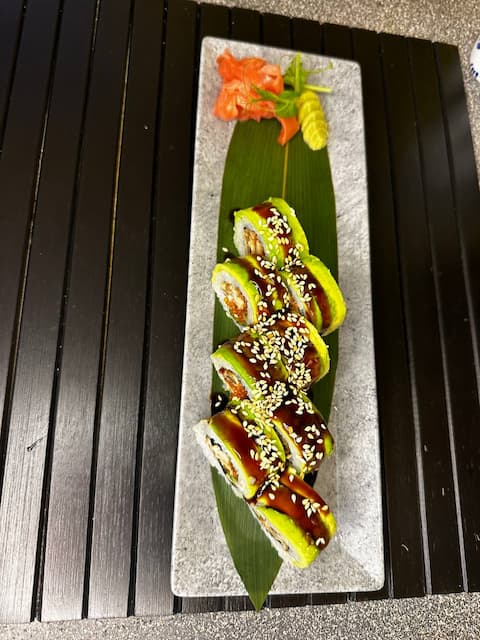 Sushi rolls with sesame seeds served on a black plate, with ginger on the side, in front of a yellow wall with decorative items