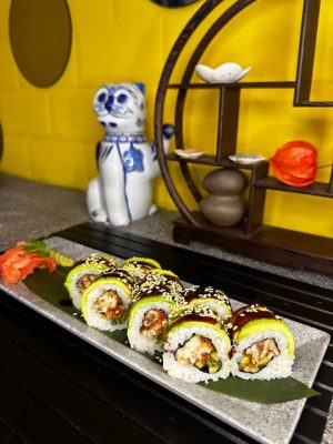 Plate of sushi rolls topped with avocado and a drizzle of sauce, garnished with sesame seeds, presented on a green leaf with chopsticks.