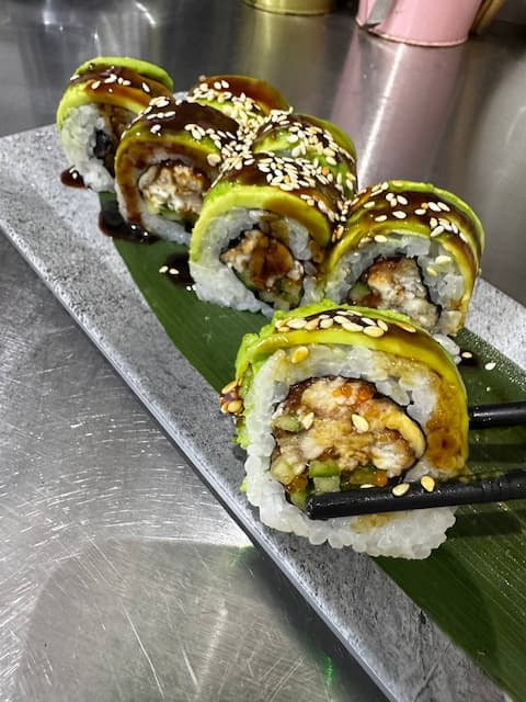 Sushi rolls with sesame seeds served on a black plate, with ginger on the side, in front of a yellow wall with decorative items.