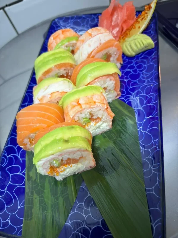 Princes Street Sushi Roll arrayed on a bamboo leaf, showcasing layers of salmon, shrimp, avocado, and mamenori, with tobiko accents, set against a blue ceramic plate background.