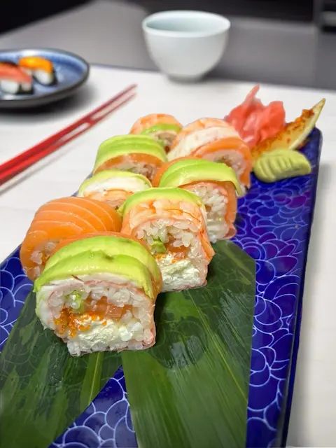 Princes Street Sushi Roll featuring shrimp, salmon, avocado, and soft cheese, wrapped in mamenori, garnished with tobiko, served on a blue textured plate with ginger and wasabi.