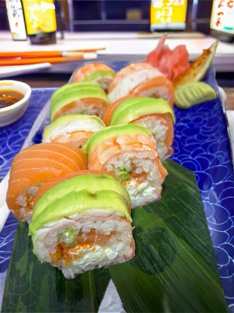 Close-up of Princes Street Sushi Roll with avocado and salmon on top, mamenori wrap, filled with shrimp, soft cheese, and tobiko, presented on a leaf over a blue plate.