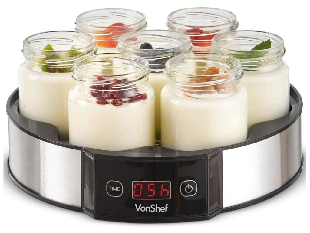 A yogurt maker with six glass jars of yogurt, each topped with different fruits or leaves, displayed on a countertop. The digital timer shows "056".